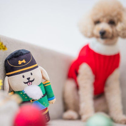Small poodle sitting beside the Woof² Nutcracker Treat-Dispensing Dog Toy, creating a festive holiday scene with blurred decorations in the foreground.