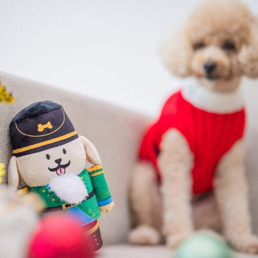 Small poodle sitting beside the Woof² Nutcracker Treat-Dispensing Dog Toy, creating a festive holiday scene with blurred decorations in the foreground.